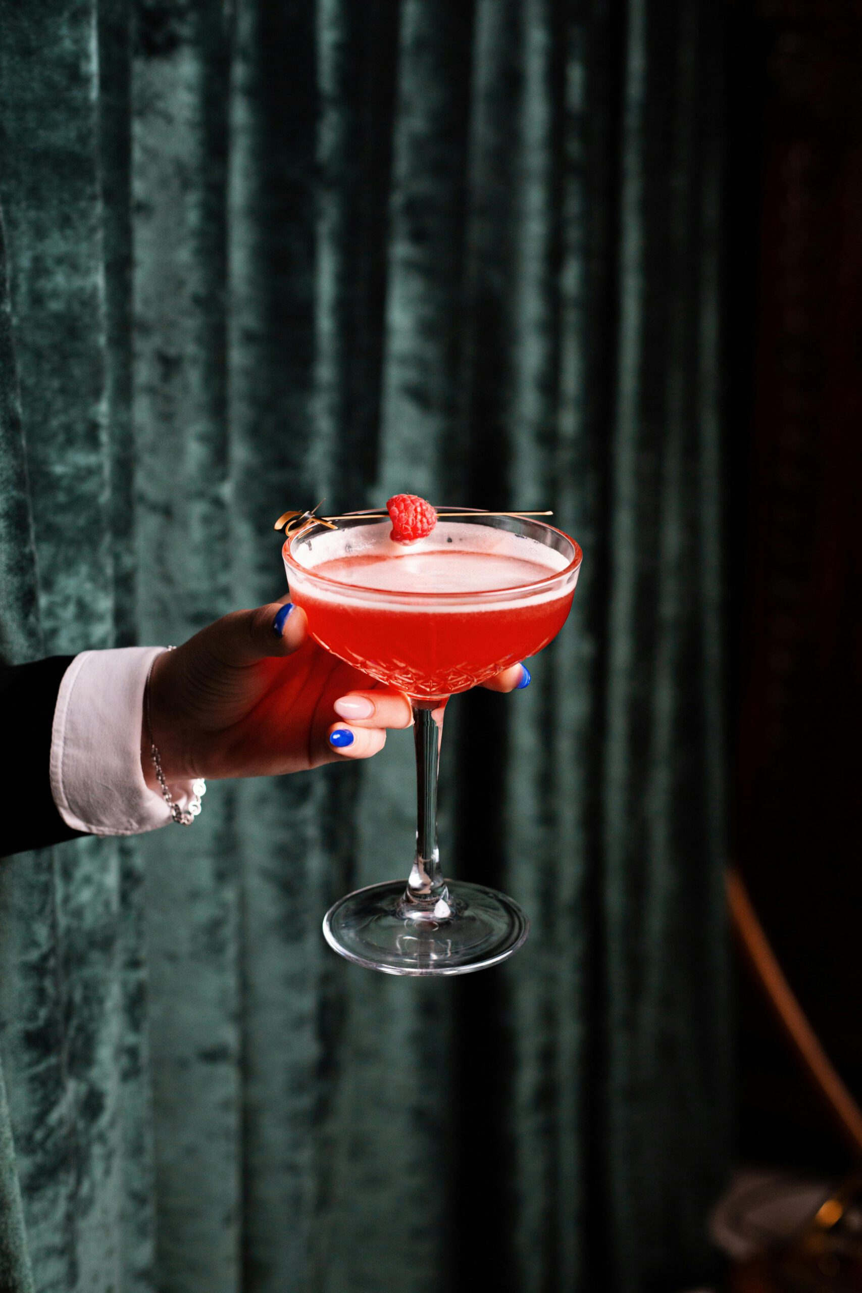 A woman's hand elegantly holding a raspberry cocktail glass against a velvet backdrop.